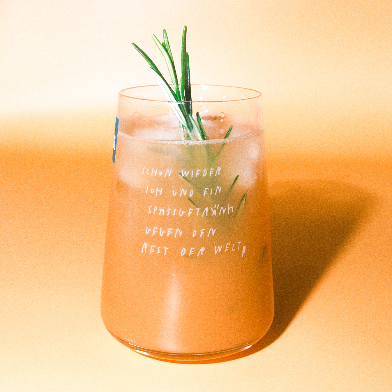 Konisches Glas mit eisgekühltem orangefarbenen Drink und Rosmarinzweig; weißer Aufdruck: „schon wieder ich und ein spaßgetränk gegen den rest der welt“.