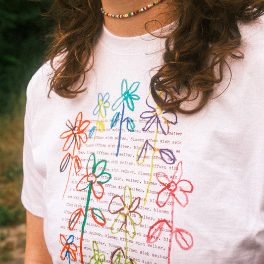 Halbnahe Frontansicht: weißes T-Shirt, mehrfarbiger Blumenprint über typografischem Hintergrund.