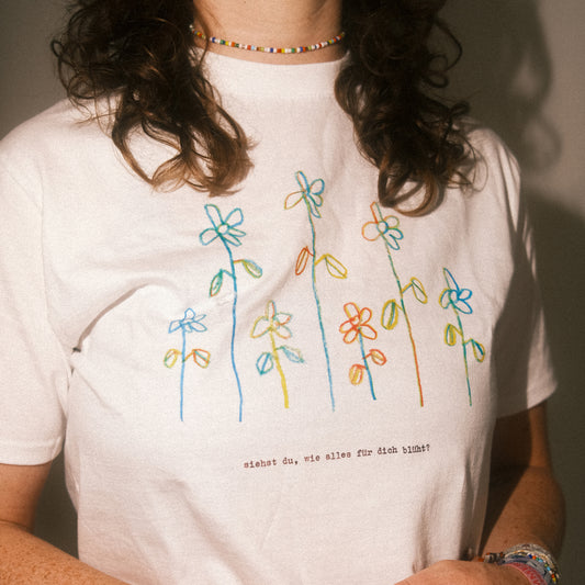 Detailaufnahme des Brustprints: bunte gezeichnete Blumenreihen; darunter „siehst du, wie alles für dich blüht?“.