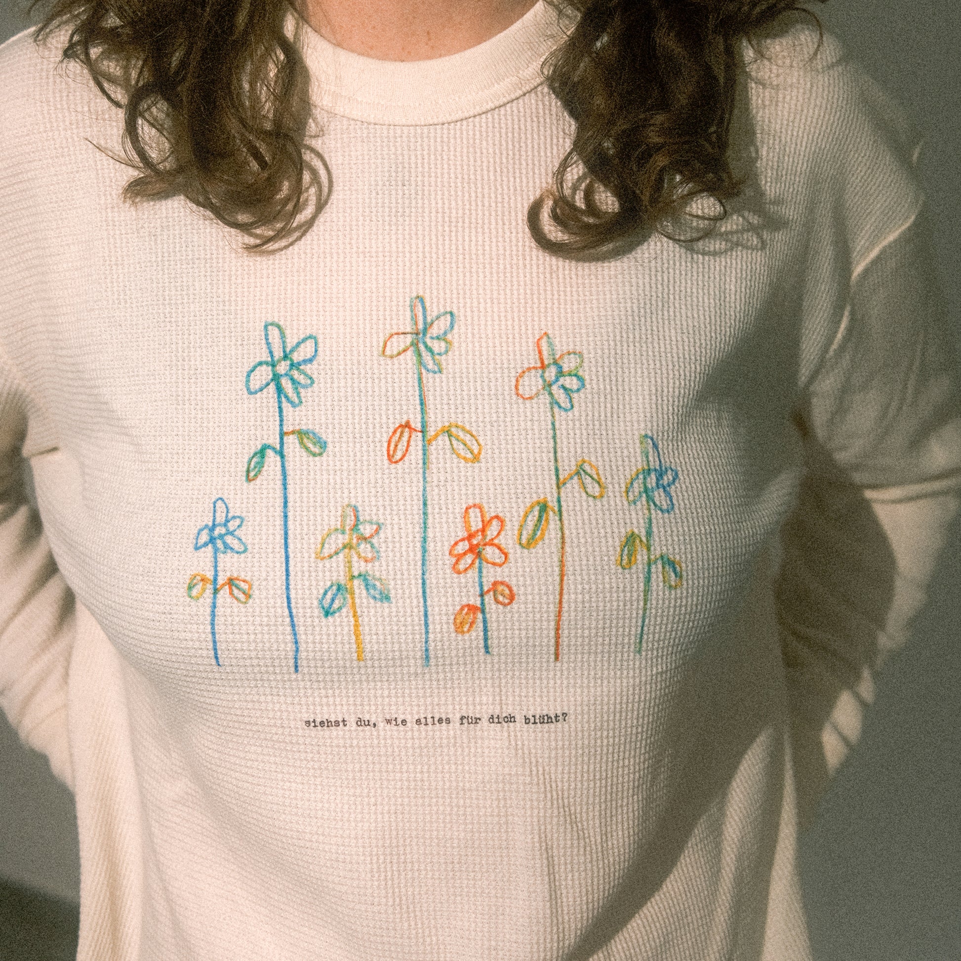 Detail Brustprint: bunte, skizzierte Blumenreihe über dem Text „siehst du, wie alles für dich blüht?“ auf Waffelstrick.