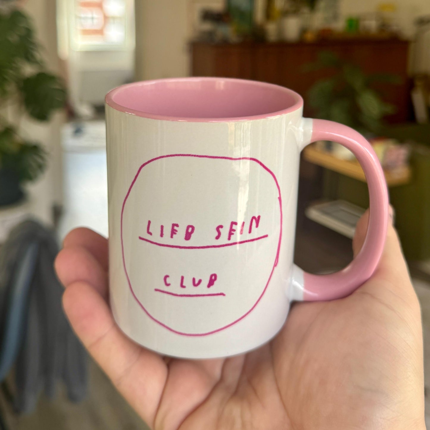 Weiße Keramiktasse mit pinkfarbenem Henkel und Innenrand. Auf der Vorderseite steht handschriftlich in pinkfarbenem Schriftzug innerhalb eines grob gezeichneten Kreises: „LIEB SEIN CLUB“.