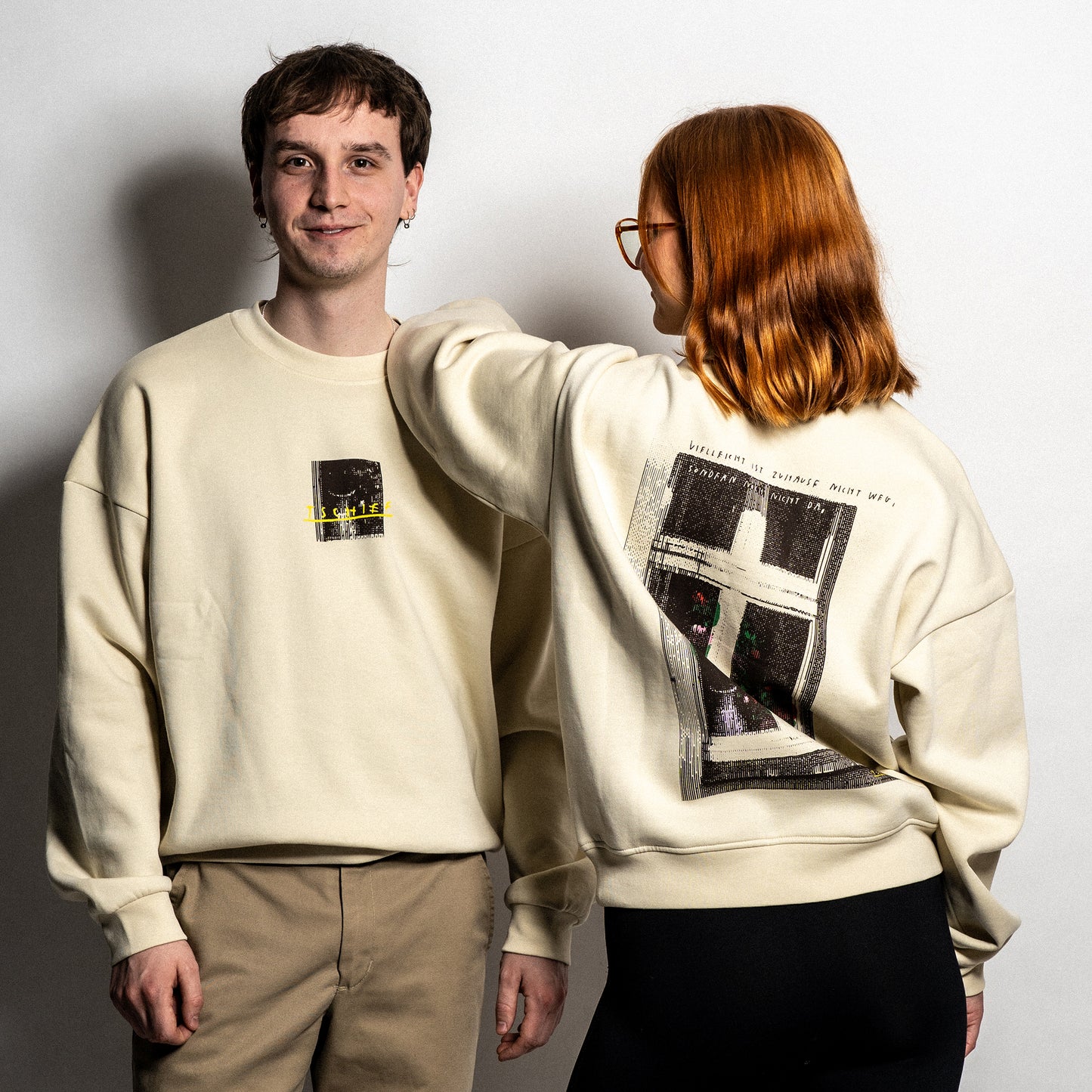 Zwei Personen tragen einen beigefarbenen Unisex-Sweater. Die linke Person zeigt die Vorderseite mit einem kleinen Frontprint: ein dunkles Fensterbild mit gelber, handschriftlicher Aufschrift „TSCHIEF“. Die rechte Person ist rückwärts gedreht und zeigt den großflächigen Rückendruck: eine stilisierte Fensterillustration mit floralen Elementen, darüber der Satz „Vielleicht ist Zuhause nicht weg, sondern nur nicht da“ in handschriftlicher Schrift.