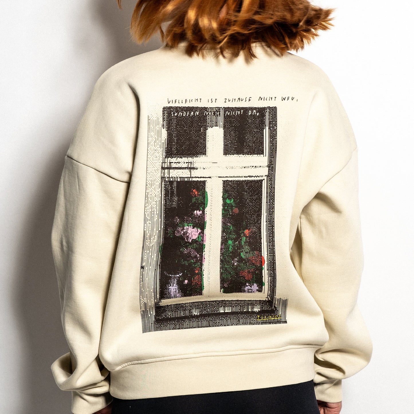 Beigefarbener Sweater mit großem Rückendruck. Das Motiv zeigt ein grafisch interpretiertes Fenster mit Blumen im unteren Bereich. Oberhalb des Bildes steht in Handschrift: „Vielleicht ist Zuhause nicht weg, sondern nur nicht da“. Das Design wirkt poetisch und nostalgisch.