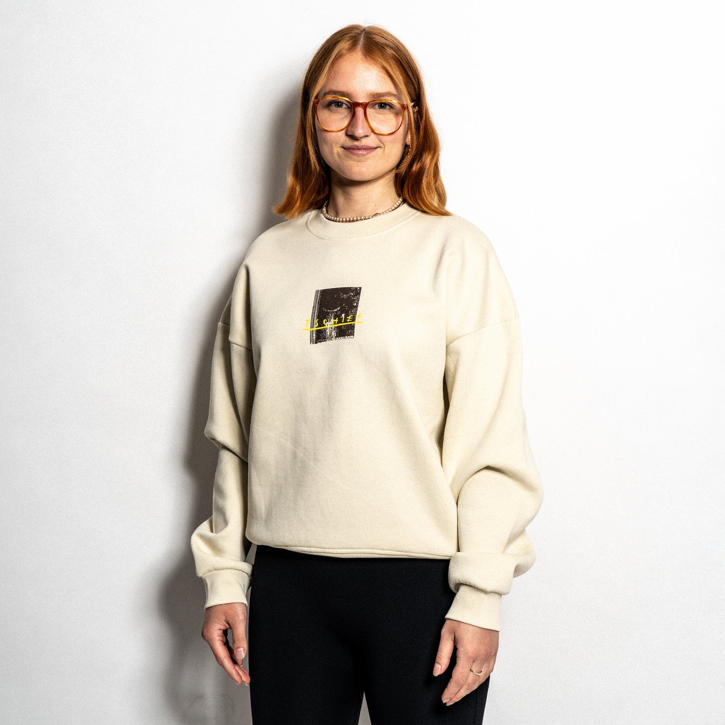 Person trägt einen beigefarbenen Sweater mit kleinem zentralem Frontprint. Zu sehen ist ein dunkel hinterlegtes Bild mit Fensterstruktur und gelb eingesticktem Schriftzug „TSCHIEF“ in handschriftlichem Stil. Der Sweater hat einen lockeren Schnitt mit tief angesetzten Schultern.