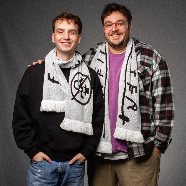 Zwei Personen tragen den doppelseitigen Winter-Schal jeweils um den Hals. Zu sehen ist einmal das Schneemann-Symbol und einmal der handschriftliche Schriftzug mit weißen Fransen an den Schalenden.