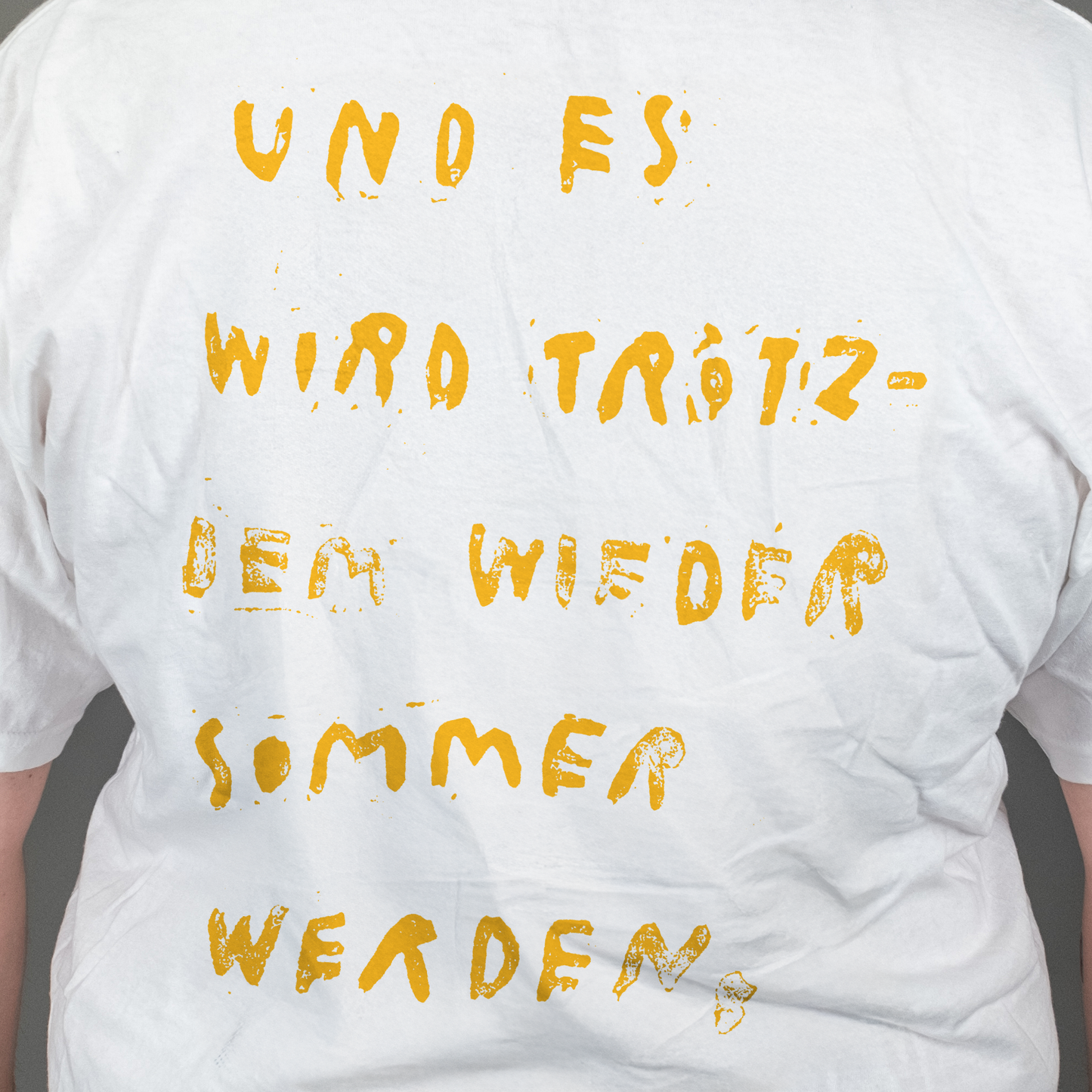 Nahaufnahme des gelben Rückendrucks auf weißem T-Shirt. Der Text in handschriftlicher, leicht ausgefranster Typografie lautet: „UND ES WIRD TRÖTZDEM WIEDER SOMMER WERDEN,“ – verteilt auf sechs Zeilen.