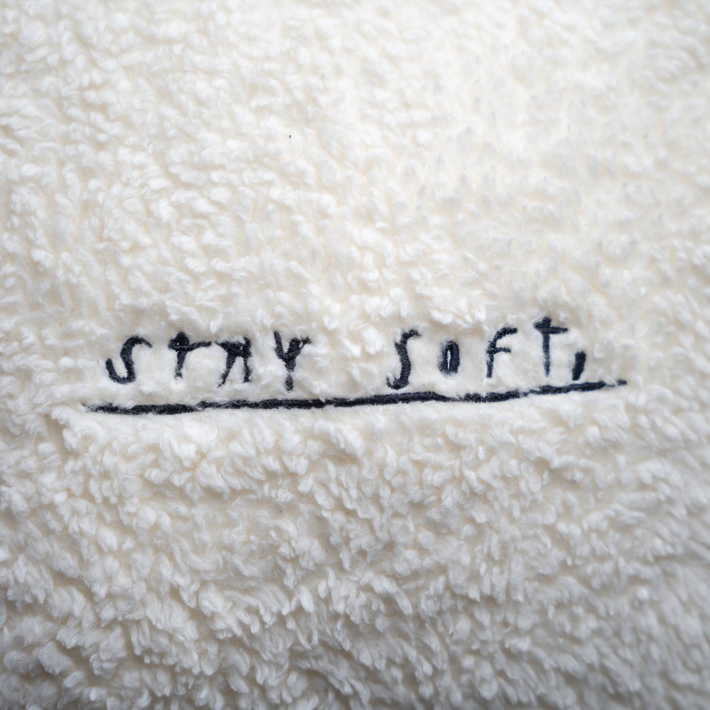 Detailansicht der Kissenoberfläche mit schwarzer Stickerei „STAY SOFT,“ in handschriftlicher Schreibweise auf dem weichen, cremeweißen Plüschstoff.