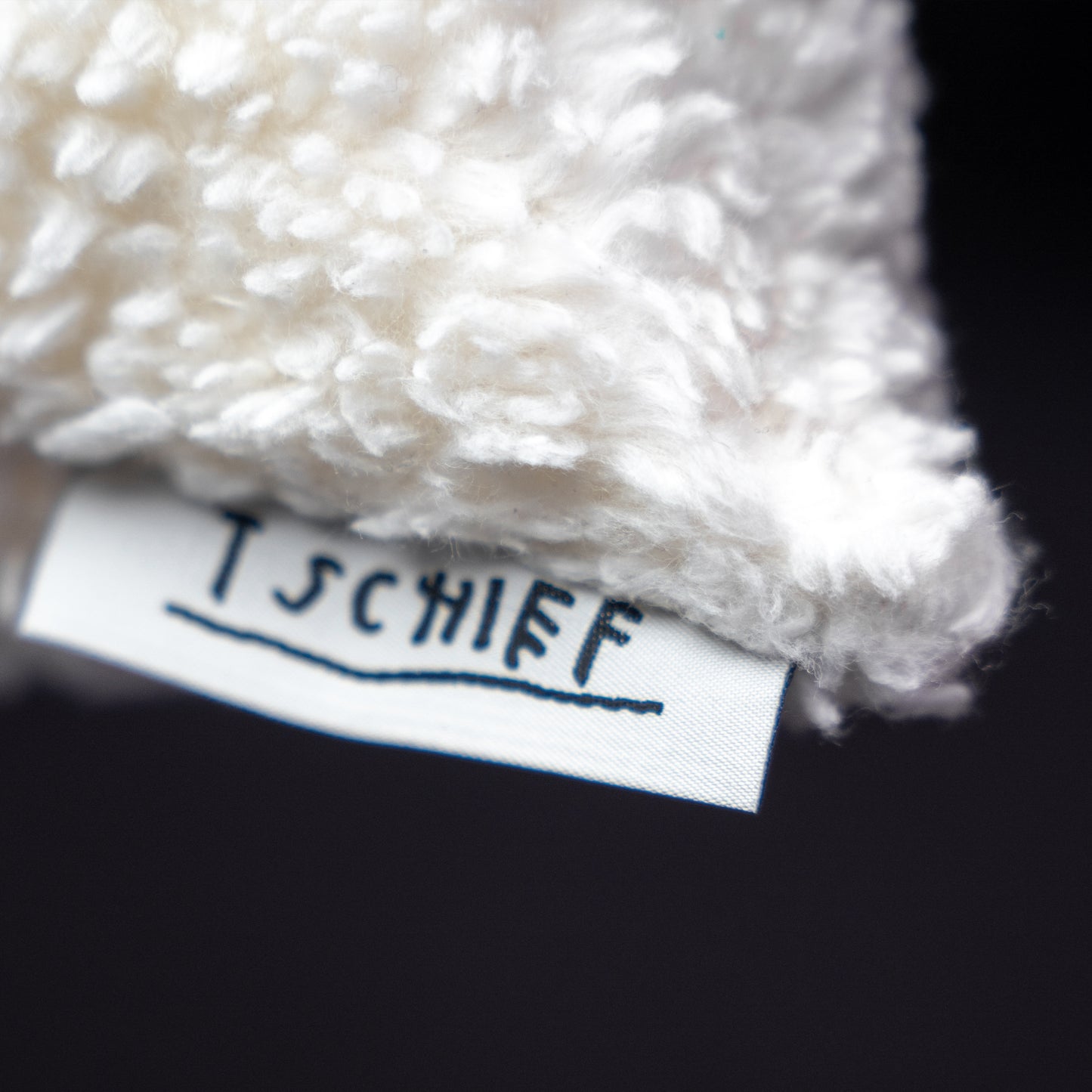 Makroaufnahme des Markenlabels „TSCHIEF“, das seitlich aus dem weißen, flauschigen Kissen hervorsteht. Der Schriftzug ist handgeschrieben und unterstrichen.