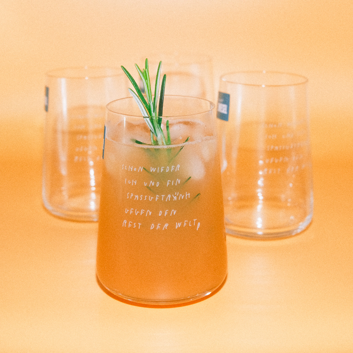 Gefülltes Glas mit Eis und Rosmarin im Vordergrund, zwei leere Gläser mit gleichem Schriftaufdruck im Hintergrund; orangener Hintergrund.