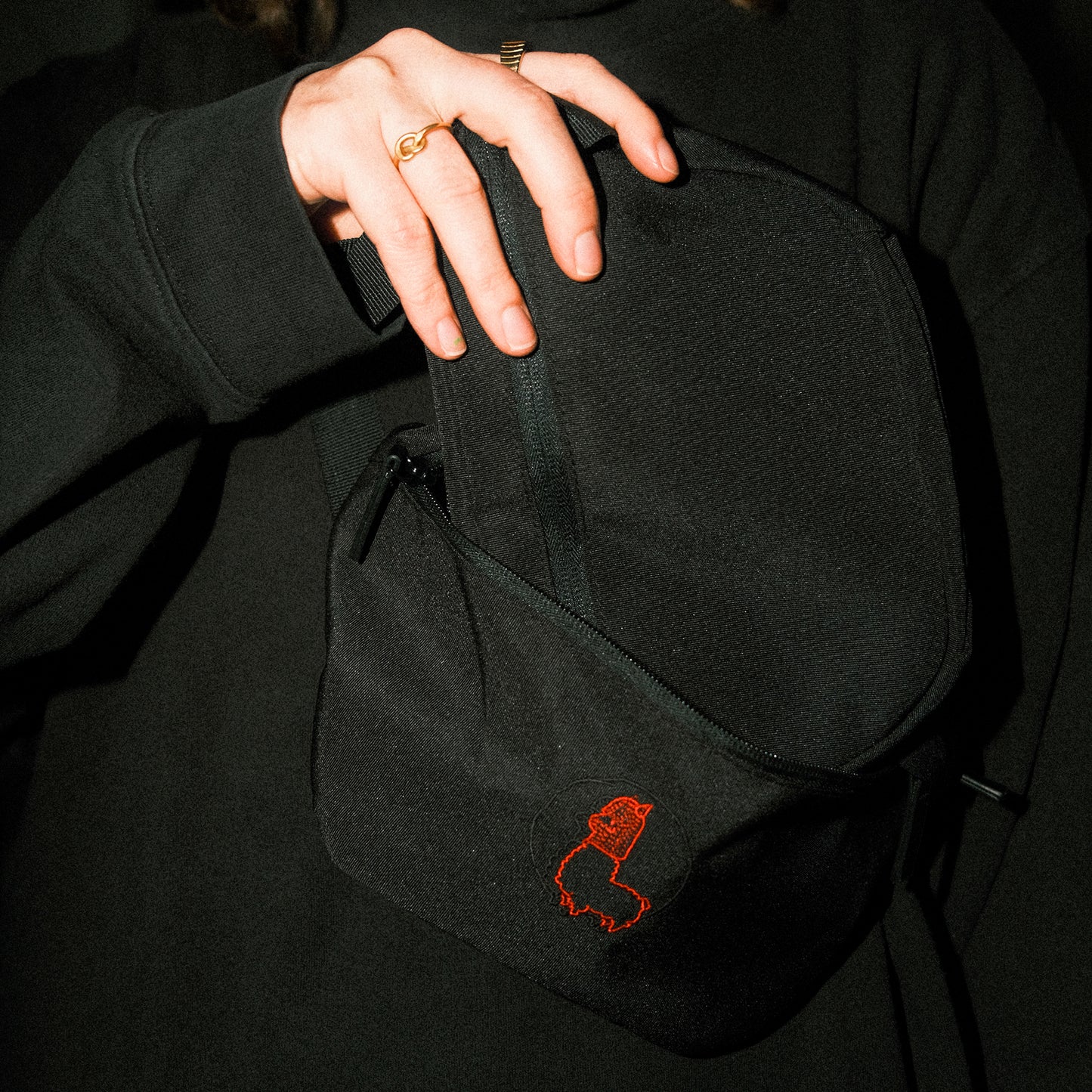 Schwarze Umhängetasche aus robustem Stoff mit Reißverschluss. Auf der Vorderseite ist ein rotes, feingesticktes Motiv eines stilisierten Alpakas in seitlicher Ansicht. Die Tasche ist geöffnet, eine Hand greift nach dem oberen Griff.
