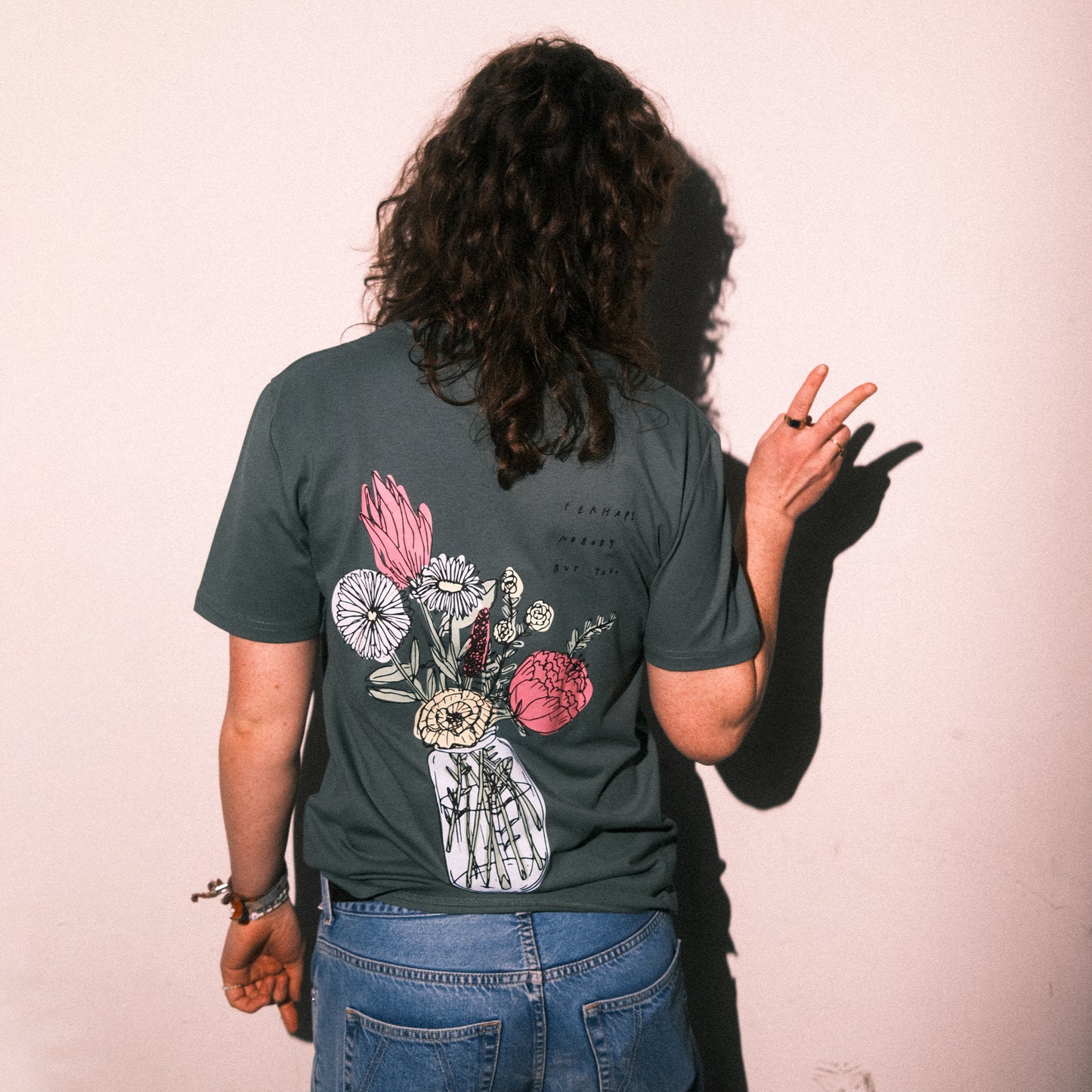 Grünes T-Shirt mit kurzen Ärmeln. Auf dem Rücken ist ein großes, farbenfrohes Blumenmotiv in einem Glas dargestellt. Daneben steht handschriftlich in schwarzer Schrift der Text: „perhaps nobody but you“. Die Person trägt das T-Shirt locker mit Jeans.