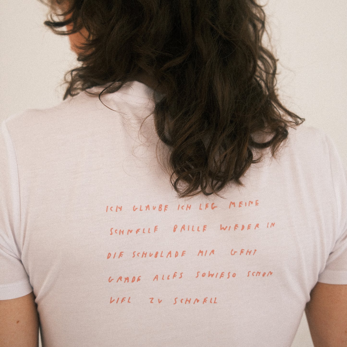 Weißes T-Shirt mit einem poetischen Textdruck auf dem Rücken. Der Text ist in locker handgeschriebener roter Schrift gehalten und lautet: „Ich glaub ich leg meine schnelle Brille wieder in die Schublade mir geht grade alles sowieso schon viel zu schnell“. Das T-Shirt hat kurze Ärmel und einen klassischen Rundhalsausschnitt. Das Material wirkt weich und leicht transparent. Die Person trägt das Shirt locker, die Haare sind leicht gewellt und die Aufnahme zeigt den Rücken. Der Hintergrund ist hell und neutral.