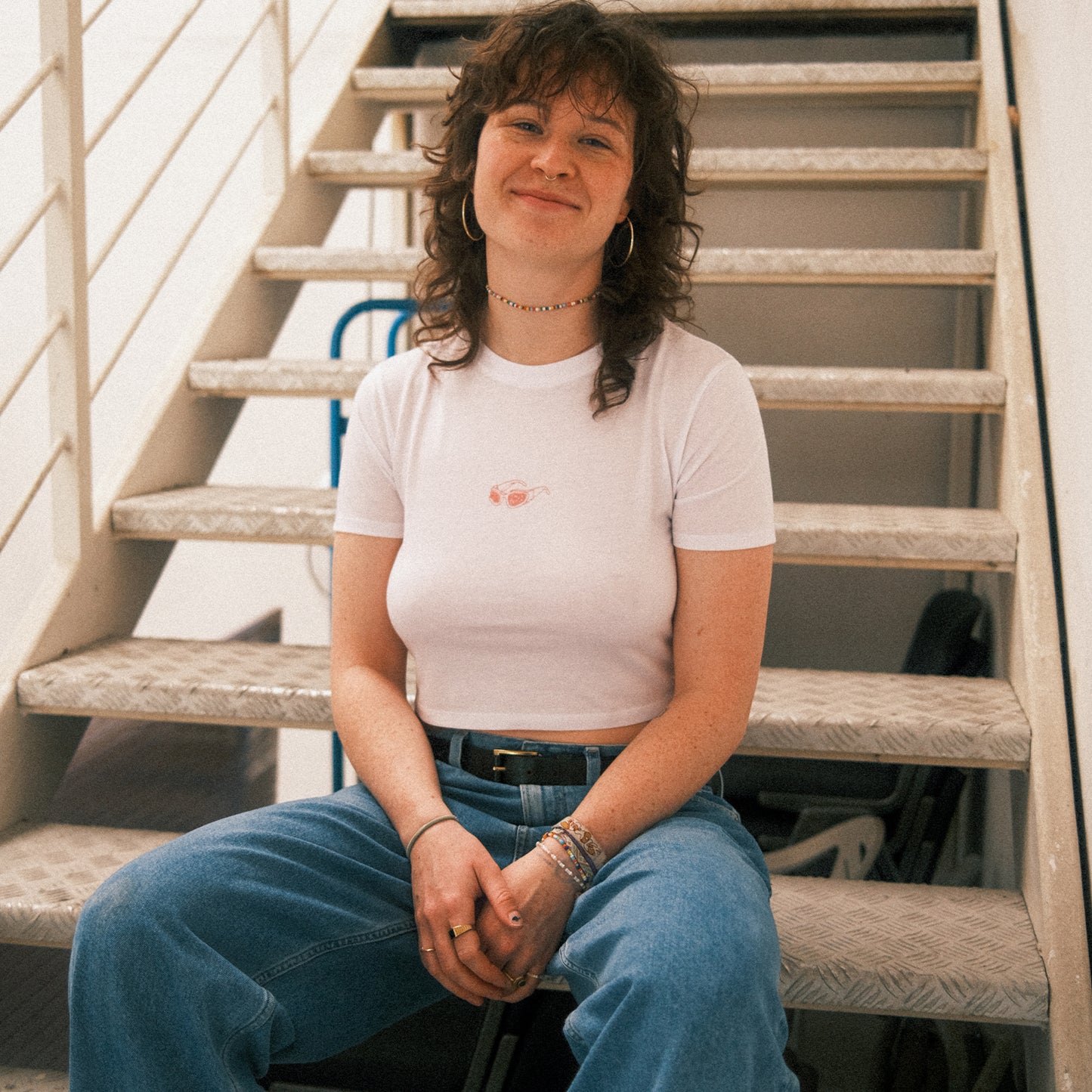 Weißes, eng anliegendes Cropped-T-Shirt mit kurzen Ärmeln. Auf der Brustmitte ist ein kleines Motiv einer rosa Sonnenbrille mit Herzgläsern abgebildet. Das T-Shirt ist schlicht und clean gestaltet, der Fokus liegt klar auf dem dezenten Brustdruck.