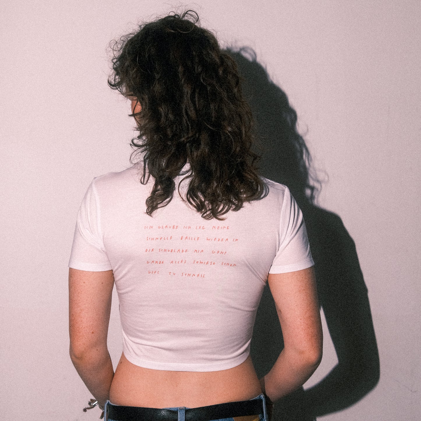 Weißes, kurzes Cropped-T-Shirt mit kurzem Ärmel. Auf dem Rücken befindet sich ein handschriftlicher, rot-orangener Text, der in mehreren Zeilen geschrieben ist: „Ich glaube ich leg meine schnelle Brille wieder in die Schublade mir geht grade alles sowieso schon viel zu schnell“. Die Person trägt das T-Shirt mit blauer Jeans und steht mit dem Rücken zur Kamera. Das Haar ist lockig und schulterlang.