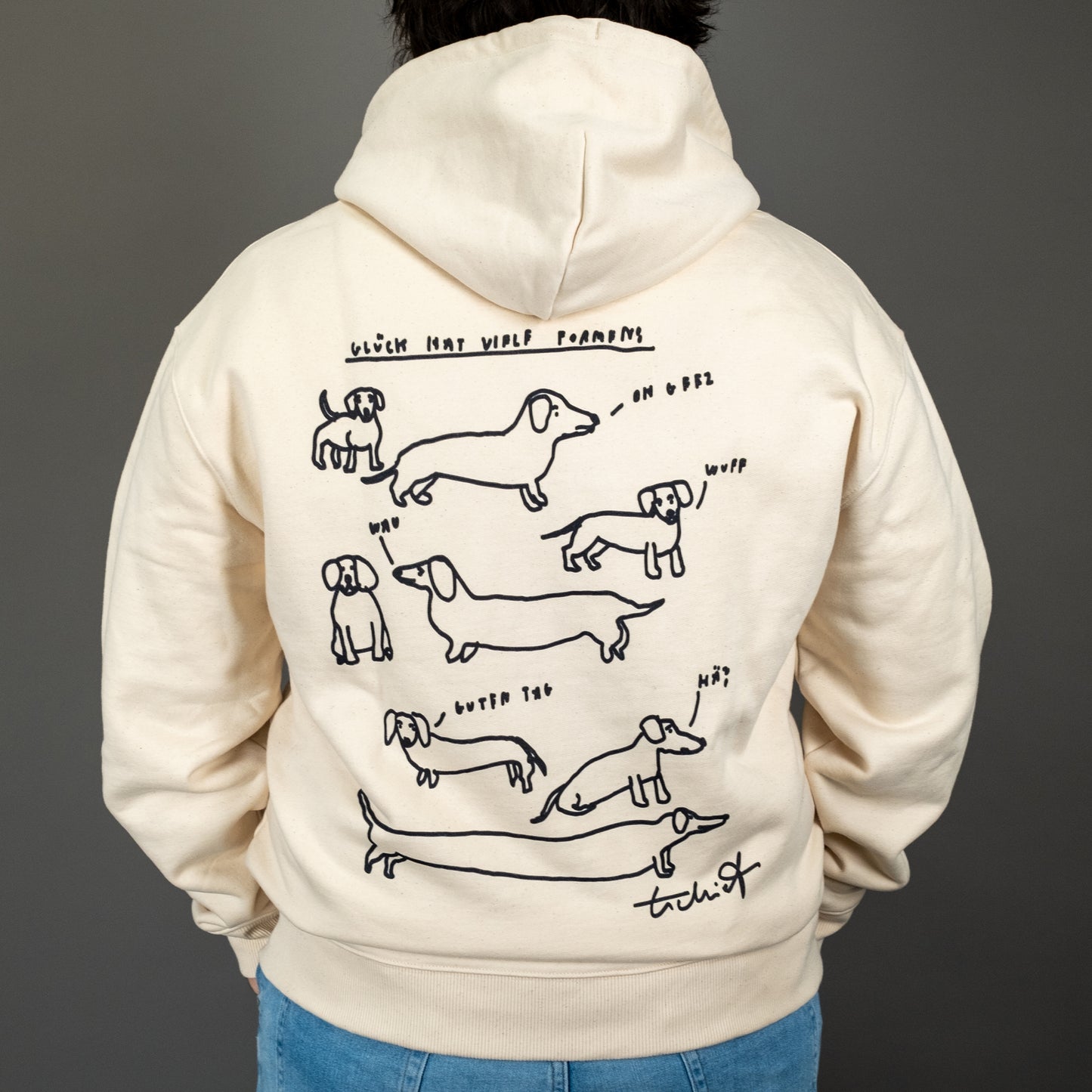 Rückenansicht eines cremefarbenen Kapuzen-Zipper-Hoodies mit schwarzem Aufdruck. Der Aufdruck zeigt mehrere einfache, gezeichnete Dackel in unterschiedlichen Größen und Posen, mit kleinen Sprechblasen, die verschiedene Laute wie „Wuff“, „Oh“, „Miau?“ und „Hä?“ enthalten. Über den Zeichnungen steht der Schriftzug „Glück hat viele Formen!“ in handschriftlicher schwarzer Schrift. Unten rechts befindet sich eine Unterschrift „Tschiff“.