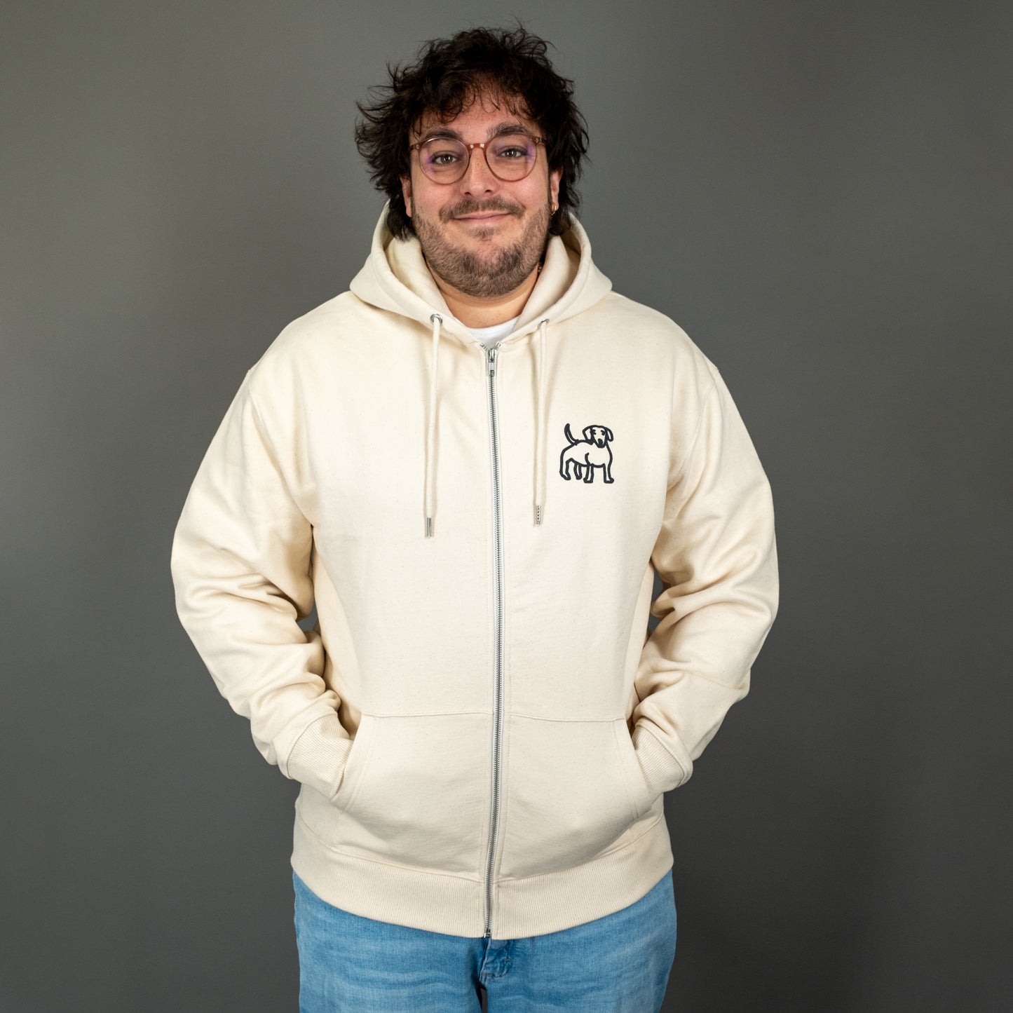 Frontalansicht eines cremefarbenen Kapuzen-Zipper-Hoodies. Auf der linken Brustseite ist ein kleiner, schwarz gezeichneter Dackel aufgestickt. Der Hoodie hat Kängurutaschen, einen Reißverschluss und weiße Kordeln an der Kapuze. Das Modell trägt den Hoodie locker und hat die Hände in den Taschen.