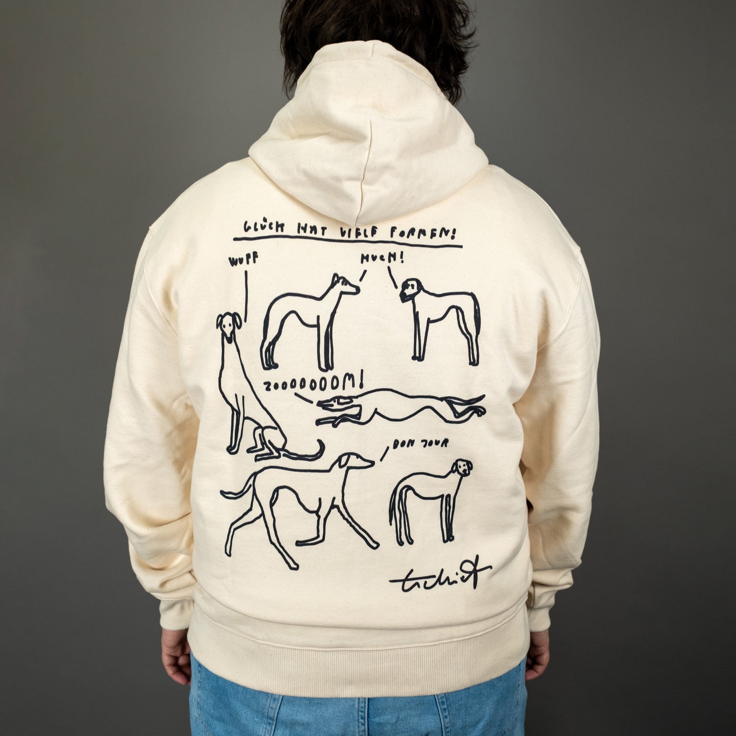 Eine Person trägt einen beigen Kapuzenpullover mit schwarzem Aufdruck auf dem Rücken. Der Aufdruck zeigt sechs handgezeichnete Hunde in verschiedenen Posen, umgeben von kleinen Sprechblasen mit den Lauten „Wuff“, „Muen!“, „Zoooooom!“ und „Bon Jour“. Darüber steht in handschriftlicher Schrift „Glück hat viele Formen!“. Unten rechts befindet sich die Signatur „tschiff“.