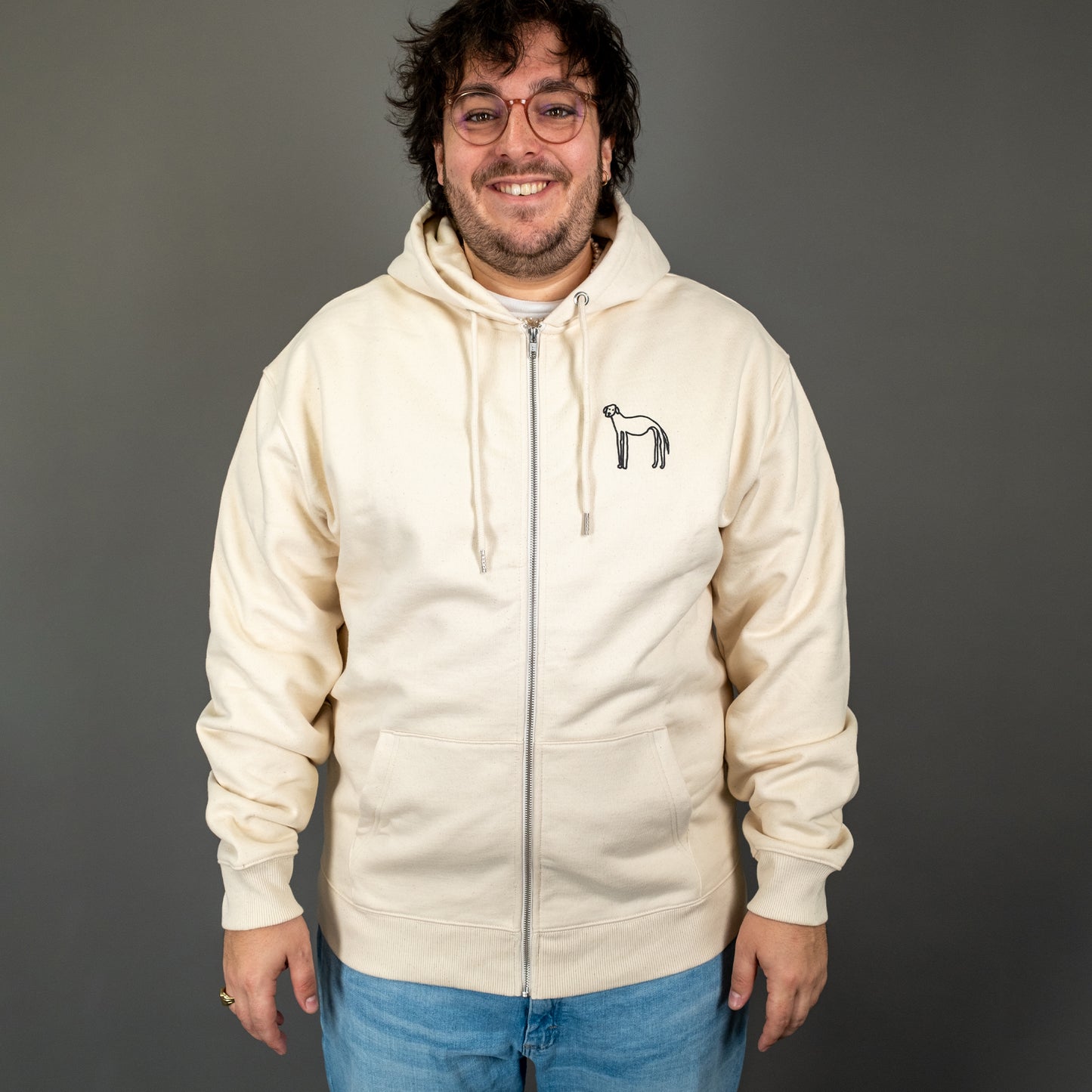 Person trägt einen beigen Kapuzenpullover mit Reißverschluss (Zipper). Auf der linken Brustseite ist ein kleiner, handgezeichneter Hund in schwarzer Farbe aufgedruckt. Der Pullover hat lange Ärmel, eine Kängurutasche und eine Kapuze mit Kordeln.