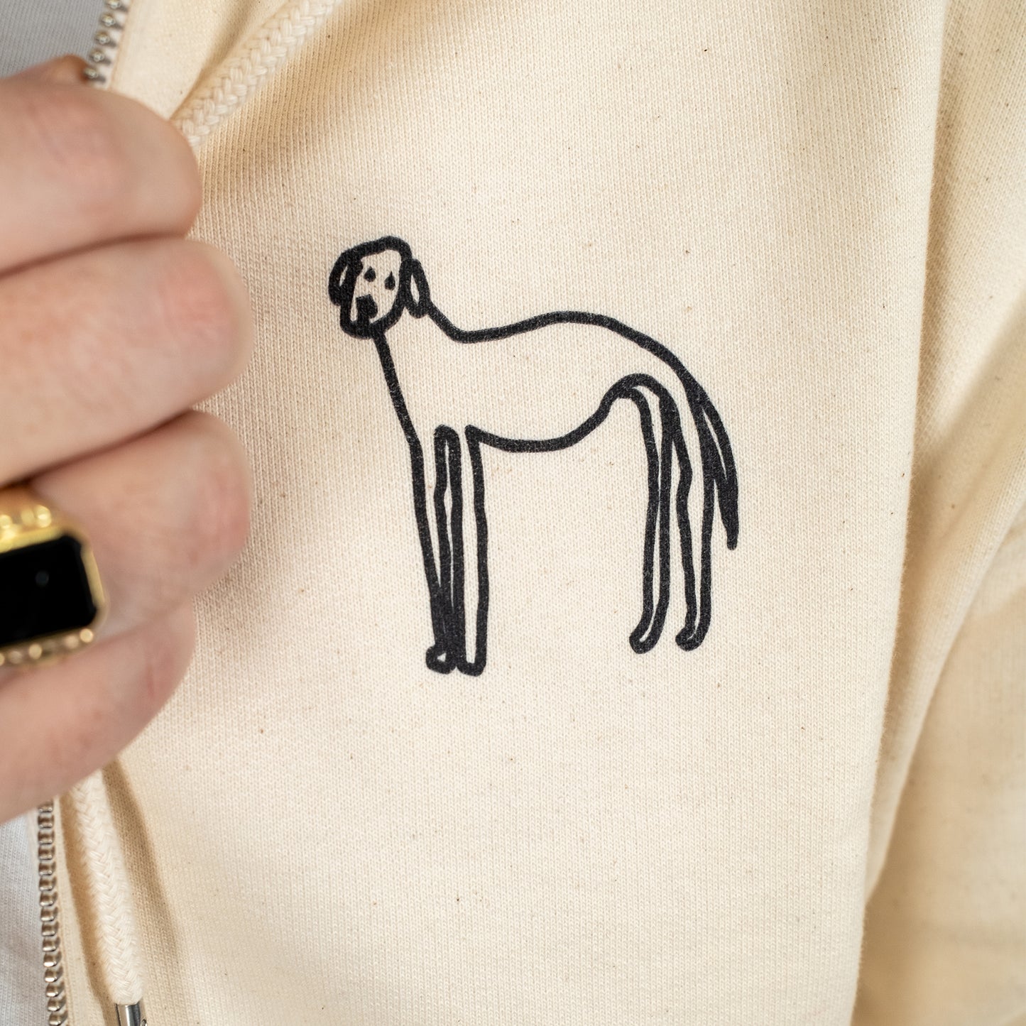 Detailaufnahme eines beigen Kapuzenpullovers mit Reißverschluss. Im Fokus steht ein handgezeichneter Hund in schwarzer Farbe auf der linken Brustseite des Pullovers. Die Zeichnung ist einfach und skizzenhaft gehalten.