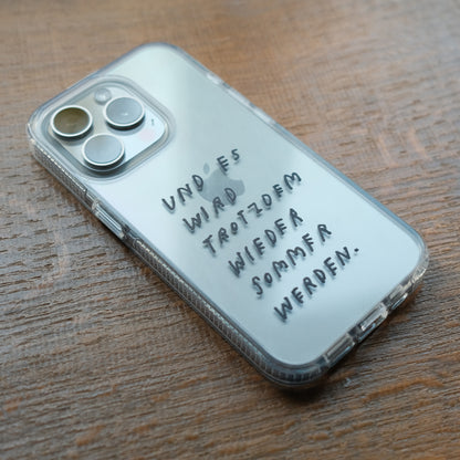 Nahaufnahme einer transparenten Handyhülle mit schwarzer Handschrift. Auf der Hülle steht:
„Und es wird trotzdem wieder Sommer werden.“