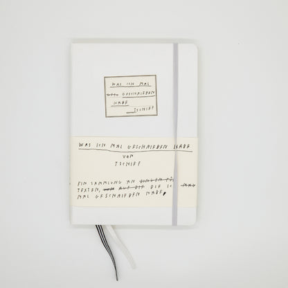 Weiße Notizbuchhülle mit einem weißen Papierband umwickelt, das handschriftlichen schwarzen Text zeigt. Auf dem Papierband steht: „WAS ICH MAL GESCHRIEBEN HABE VON TSCHIFF EIN SAMMLUNG AN TEXTEN, DIE ICH MAL GESCHRIEBEN HABE“ mit einigen durchgestrichenen Wörtern. Auf der Buchhülle ist ein kleiner weißer quadratischer Aufkleber mit schwarzem Rand und handschriftlichem Text in Schwarz, der ähnlich lautet. Ein graues Gummiband zum Verschließen des Buchs ist an der rechten Seite zu sehen. Zwei Lesezeichenbände