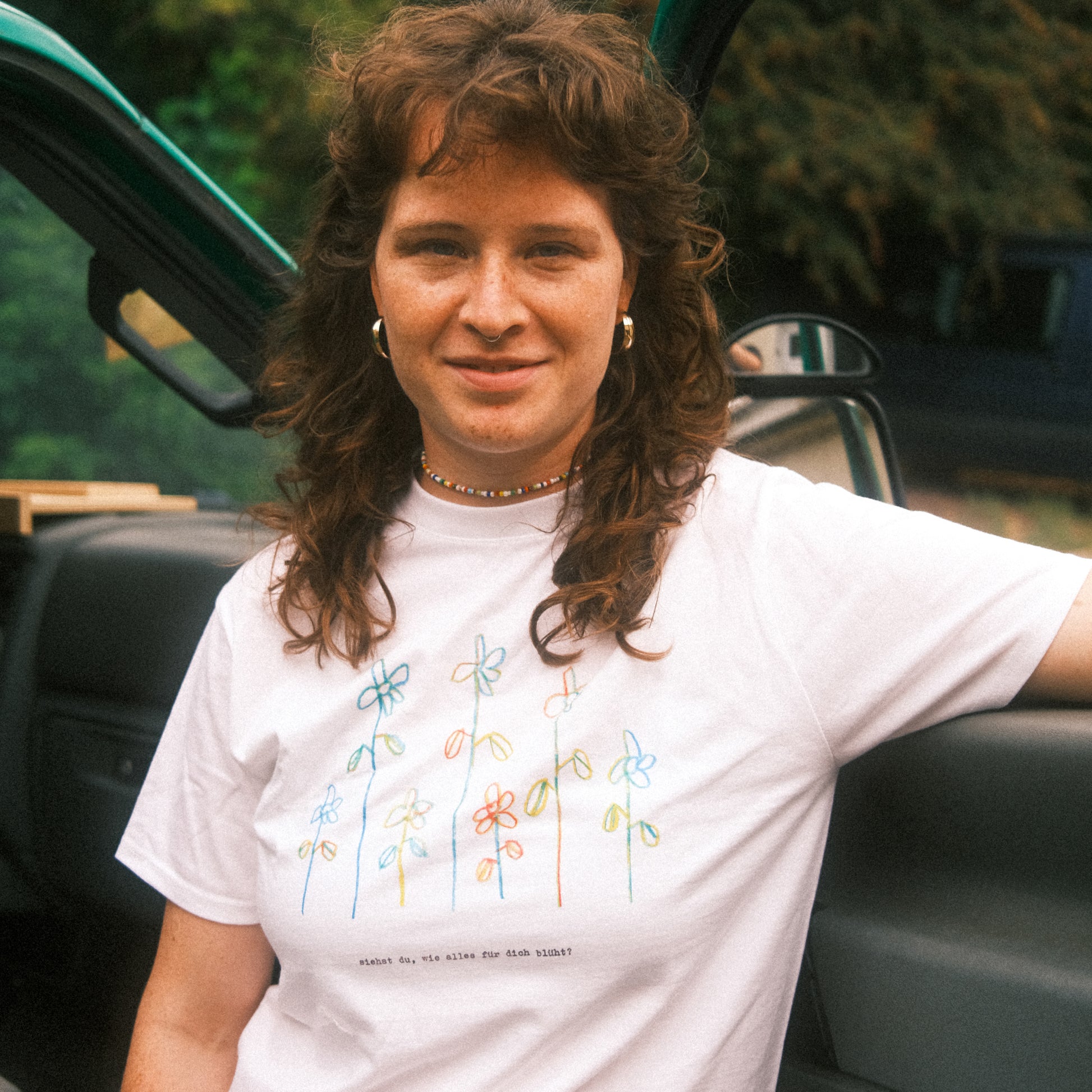 Weißes T-Shirt mit buntem Line-Art-Blumenprint und Text „siehst du, wie alles für dich blüht?“; lässig an einer Autotür stehend.
