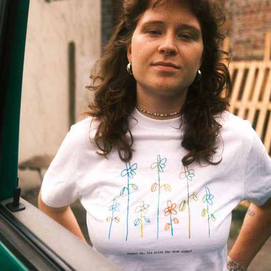 Nahaufnahme am Fahrzeugfenster: weißes T-Shirt mit bunter Blumenreihe und dem Text „siehst du, wie alles für dich blüht?“.