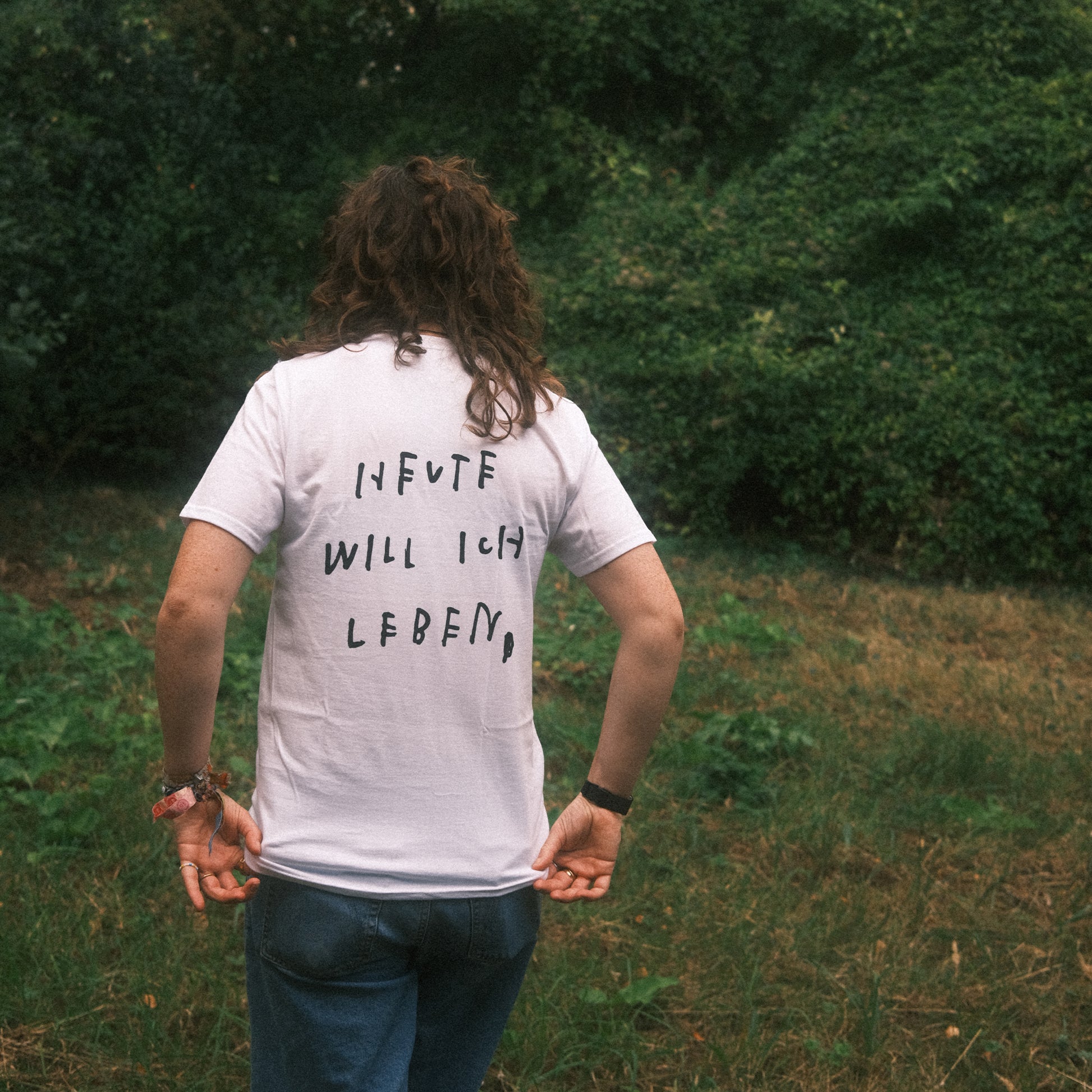 Weißes T-Shirt, Rückenansicht draußen: großer schwarzer Statement-Print „HEUTE WILL ICH LEBEN“, Person hält den Saum.