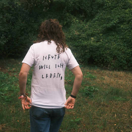Weißes T-Shirt, Rückenansicht draußen: großer schwarzer Statement-Print „HEUTE WILL ICH LEBEN“, Person hält den Saum.