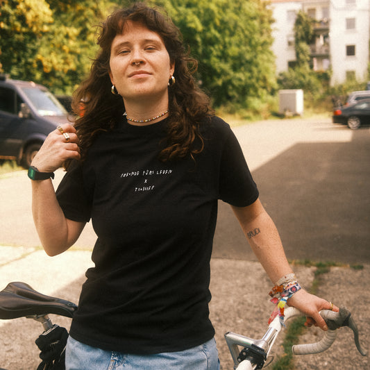 Schwarzes T-Shirt mit kleinem weißen Brustprint „FREUNDE FÜRS LEBEN × TSCHIFF“, draußen; Person hält einen Fahrradlenker.