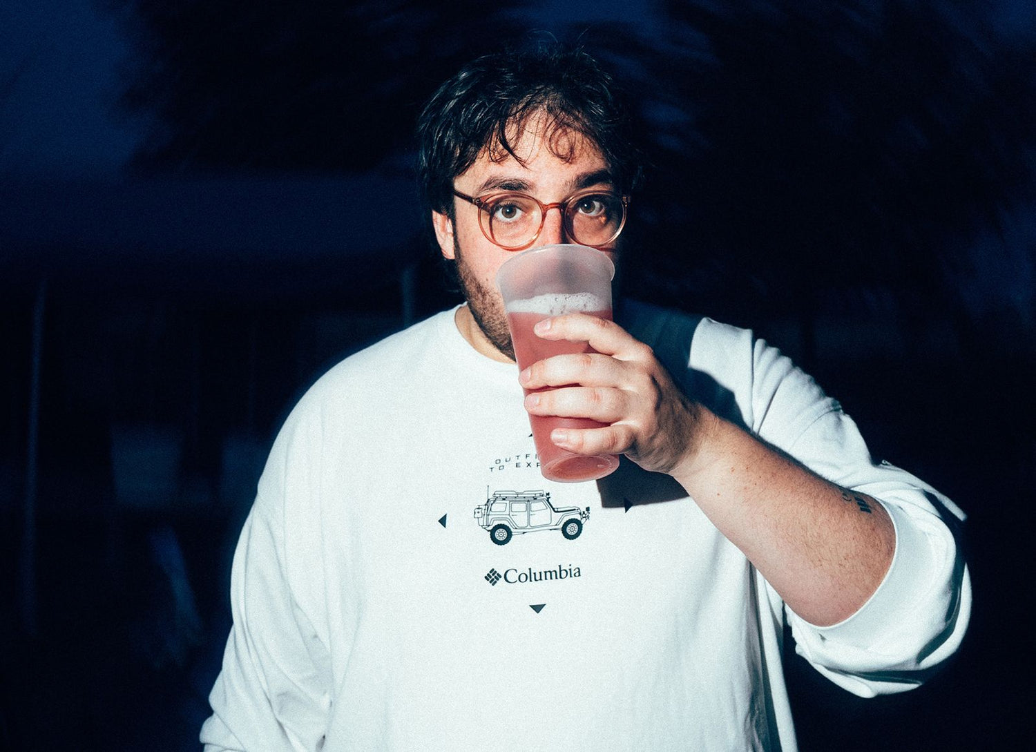 Tschief bei Nachtaufnahme mit weißem Columbia-Sweatshirt, hält einen transparenten Becher mit rosa Getränk in der Hand und blickt direkt in die Kamera. Der Hintergrund ist dunkel und unscharf, das Motiv wird von Blitzlicht beleuchtet.