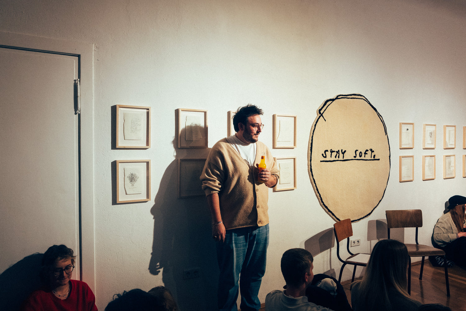 Tschief steht mit gelbem Getränk in der Hand vor einer Wand mit gerahmten Zeichnungen in einer Galerie oder Ausstellungsumgebung. Neben ihm befindet sich ein großes rundes Wandtextil mit der Aufschrift „STAY SOFT“. Im Vordergrund sitzen mehrere Personen auf dem Boden, zwei leere Stühle stehen an der Wand.
