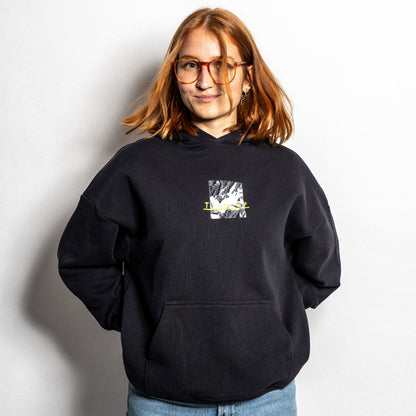 Schwarzer Hoodie mit kleinem zentralem Frontprint auf Brusthöhe. Der Druck zeigt ein fragmentiertes Schwarz-Weiß-Motiv mit grafischer Struktur und gelber Textzeile, die das Wort „I“ sowie handschriftlich anmutende Elemente beinhaltet. Der Hoodie besitzt eine Kängurutasche, breite Bündchen und eine Kapuze ohne Kordel. Der Schnitt ist lässig und unisex.