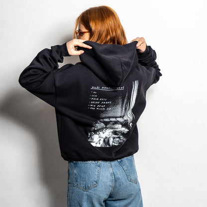 Rückansicht eines schwarzen Hoodies mit großem Rückenprint in Schwarz-Weiß. Der Druck zeigt eine fragmentarische Bildkomposition mit einem zerwühlten Bett im unteren Bereich und vertikalen Strukturen, die an eine Vorhangszene erinnern. Darüber steht in handschriftlicher Schrift: „ALLE DURCHGEKNALLT“ mit einer Aufzählung: „– DU – ICH – DEIN BETT – DEINE SORGE – WIR ZWEI – VON VORN“. Die Person trägt die Kapuze leicht angehoben und kombiniert den Hoodie mit einer hellblauen Jeans.


