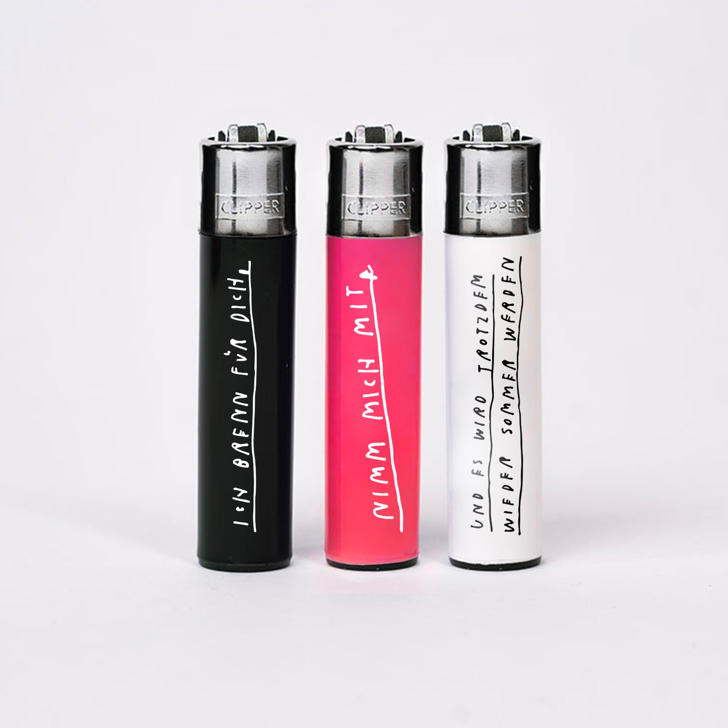 Drei Clipper-Feuerzeuge stehend auf weißem Hintergrund, jeweils mit handschriftlichen weißen oder schwarzen Texten. Das schwarze Feuerzeug links trägt den Schriftzug „Ich brenne für dich“, das pinkfarbene mittlere „Nimm mich mit“ und das weiße rechts „Und es wird trotzdem wieder Sommer werden“.