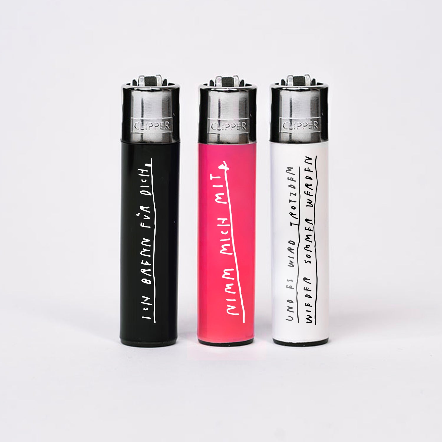 Drei Clipper-Feuerzeuge stehend auf weißem Hintergrund, jeweils mit handschriftlichen weißen oder schwarzen Texten. Das schwarze Feuerzeug links trägt den Schriftzug „Ich brenne für dich“, das pinkfarbene mittlere „Nimm mich mit“ und das weiße rechts „Und es wird trotzdem wieder Sommer werden“.