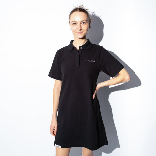 Schwarzes Polokleid mit klassischem Kragen und kurzer Knopfleiste, getragen von einer Person vor hellem Hintergrund. Auf der linken Brust ist der Schriftzug „STAY SOFT“ in Weiß aufgestickt. Das Kleid hat eine lockere, gerade Passform und reicht bis über das Knie.

