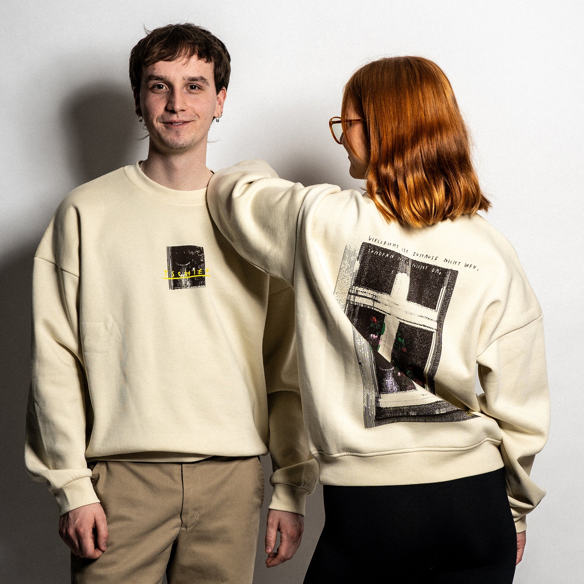 Zwei Personen tragen einen beigefarbenen Unisex-Sweater. Die linke Person zeigt die Vorderseite mit einem kleinen Frontprint: ein dunkles Fensterbild mit gelber, handschriftlicher Aufschrift „TSCHIEF“. Die rechte Person ist rückwärts gedreht und zeigt den großflächigen Rückendruck: eine stilisierte Fensterillustration mit floralen Elementen, darüber der Satz „Vielleicht ist Zuhause nicht weg, sondern nur nicht da“ in handschriftlicher Schrift.


