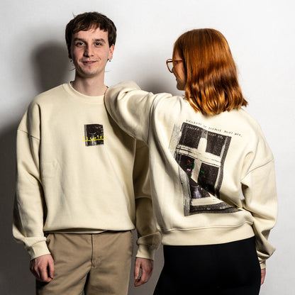 Zwei Personen tragen einen beigefarbenen Unisex-Sweater. Die linke Person zeigt die Vorderseite mit einem kleinen Frontprint: ein dunkles Fensterbild mit gelber, handschriftlicher Aufschrift „TSCHIEF“. Die rechte Person ist rückwärts gedreht und zeigt den großflächigen Rückendruck: eine stilisierte Fensterillustration mit floralen Elementen, darüber der Satz „Vielleicht ist Zuhause nicht weg, sondern nur nicht da“ in handschriftlicher Schrift.

