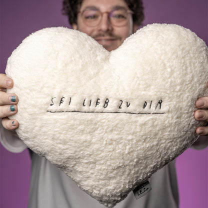 Herzförmiges Kissen aus cremeweißem, flauschigem Teddy-Stoff. In der Mitte ist der handschriftlich gestickte Schriftzug „SEI LIEB ZU DIR“ mit Unterstreichung zu sehen. Unten links am Kissenrand befindet sich ein kleines angenähtes Stofflabel mit dem Wort „LIEB“.
