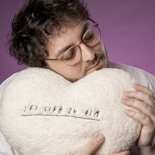 Herzförmiges, flauschiges Kissen aus cremeweißem Teddy-Stoff mit schwarzer Stickerei. In der Mitte steht in handschriftlicher Optik der Satz „SEI LIEB ZU DIR“, unterstrichen mit einer gestickten Linie.