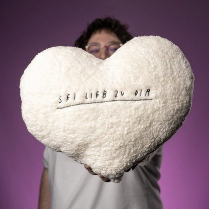 Herzförmiges, flauschiges Kissen aus cremeweißem Teddy-Stoff mit schwarzer Stickerei. In der Mitte steht in handschriftlicher Optik der Satz „SEI LIEB ZU DIR“, unterstrichen mit einer gestickten Linie.