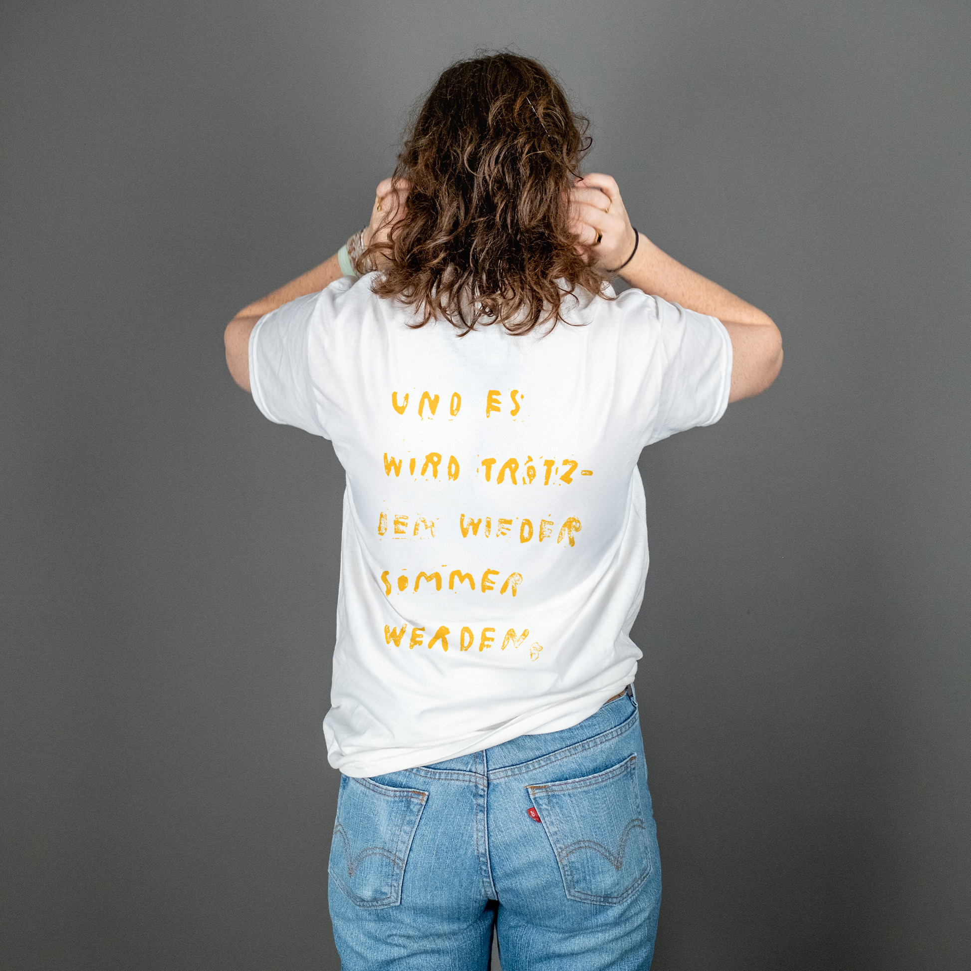 Person mit schulterlangen Locken steht mit dem Rücken zur Kamera und trägt ein weißes T-Shirt mit gelbem Rückendruck. Der handschriftlich wirkende Text lautet: „UND ES WIRD TRÖTZDEM WIEDER SOMMER WERDEN,“ – aufgeteilt in sechs Zeilen und unregelmäßig gesetzt.