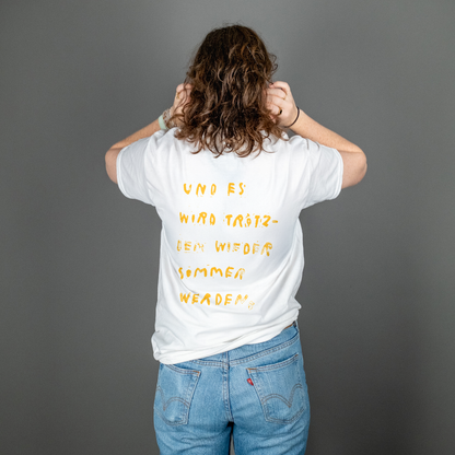Person mit schulterlangen Locken steht mit dem Rücken zur Kamera und trägt ein weißes T-Shirt mit gelbem Rückendruck. Der handschriftlich wirkende Text lautet: „UND ES WIRD TRÖTZDEM WIEDER SOMMER WERDEN,“ – aufgeteilt in sechs Zeilen und unregelmäßig gesetzt.