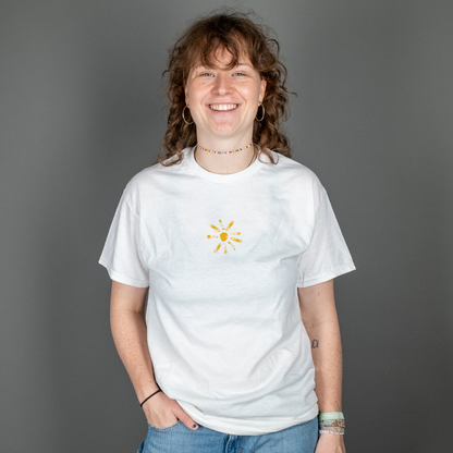 Lächelnde Person mit lockigen Haaren trägt ein weißes T-Shirt mit gelber Sonnenillustration auf der Vorderseite. Die Sonne ist mittig auf der Brust abgebildet und hat eine verspielte, gestempelte Optik.

