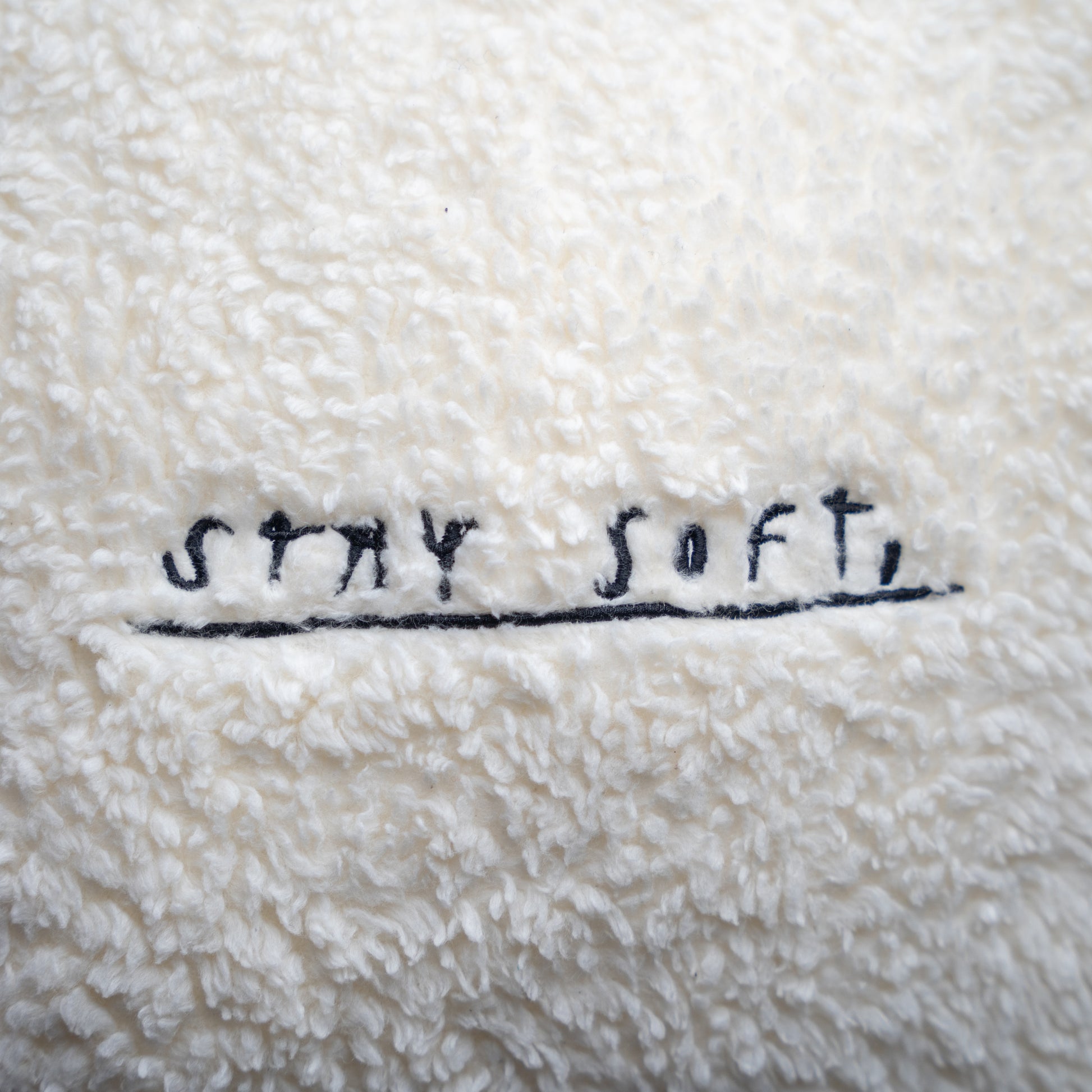 Detailansicht der Kissenoberfläche mit schwarzer Stickerei „STAY SOFT,“ in handschriftlicher Schreibweise auf dem weichen, cremeweißen Plüschstoff.