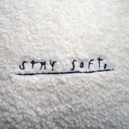 Detailansicht der Kissenoberfläche mit schwarzer Stickerei „STAY SOFT,“ in handschriftlicher Schreibweise auf dem weichen, cremeweißen Plüschstoff.