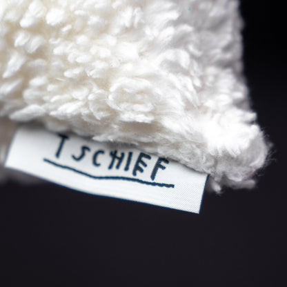 Makroaufnahme des Markenlabels „TSCHIEF“, das seitlich aus dem weißen, flauschigen Kissen hervorsteht. Der Schriftzug ist handgeschrieben und unterstrichen.