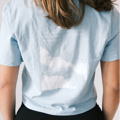 Junge Frau trägt ein hellblaues Crop-T-Shirt mit kurzem Arm, das eng anliegt. Auf der Rückseite des Shirts befindet sich ein rechteckiger Druck mit einem Bild von blauem Himmel und weißen Wolken. Darüber steht in weißer, handschriftlich anmutender Schrift ein kurzer Text, der schwer lesbar ist. Das T-Shirt liegt leicht gerafft am unteren Rücken. Die Frau trägt schwarze Hosen und steht vor einem neutralen hellen Hintergrund. Ihre braunen Haare sind schulterlang.