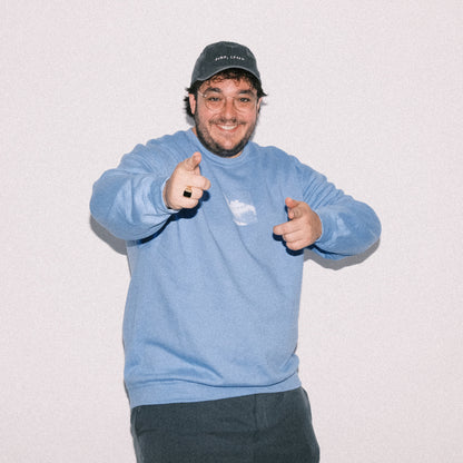 Hellblauer Sweater mit weißem Wolkenmotiv auf der Brust, getragen von einem lächelnden Mann. Der Sweater hat einen lockeren Schnitt. Der Mann trägt dazu eine dunkelblaue Cap, der Hintergrund ist hell und neutral.